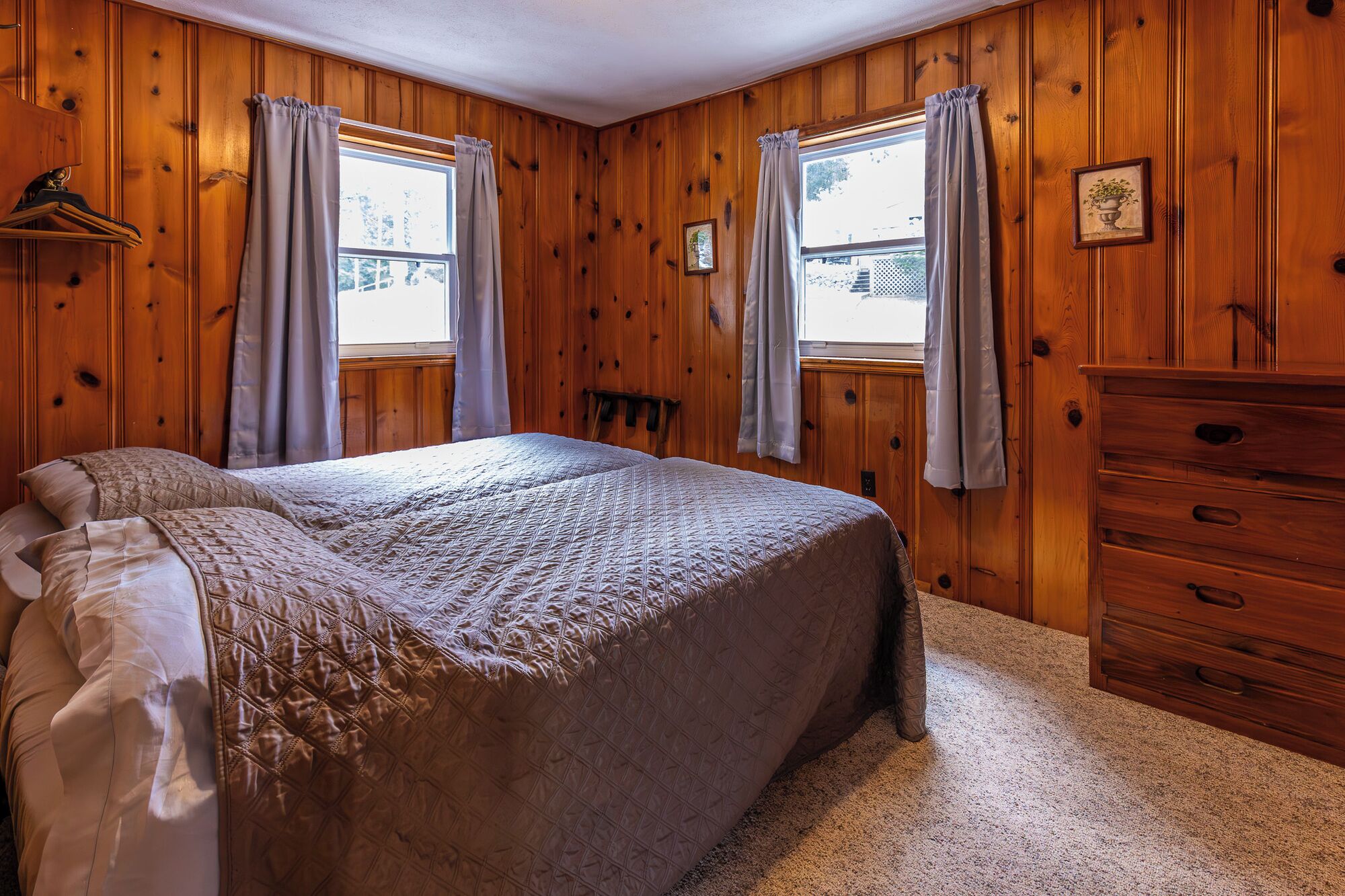 Un dormitorio con una cama grande, paredes de madera, dos ventanas con cortinas, un aparador y un perchero.