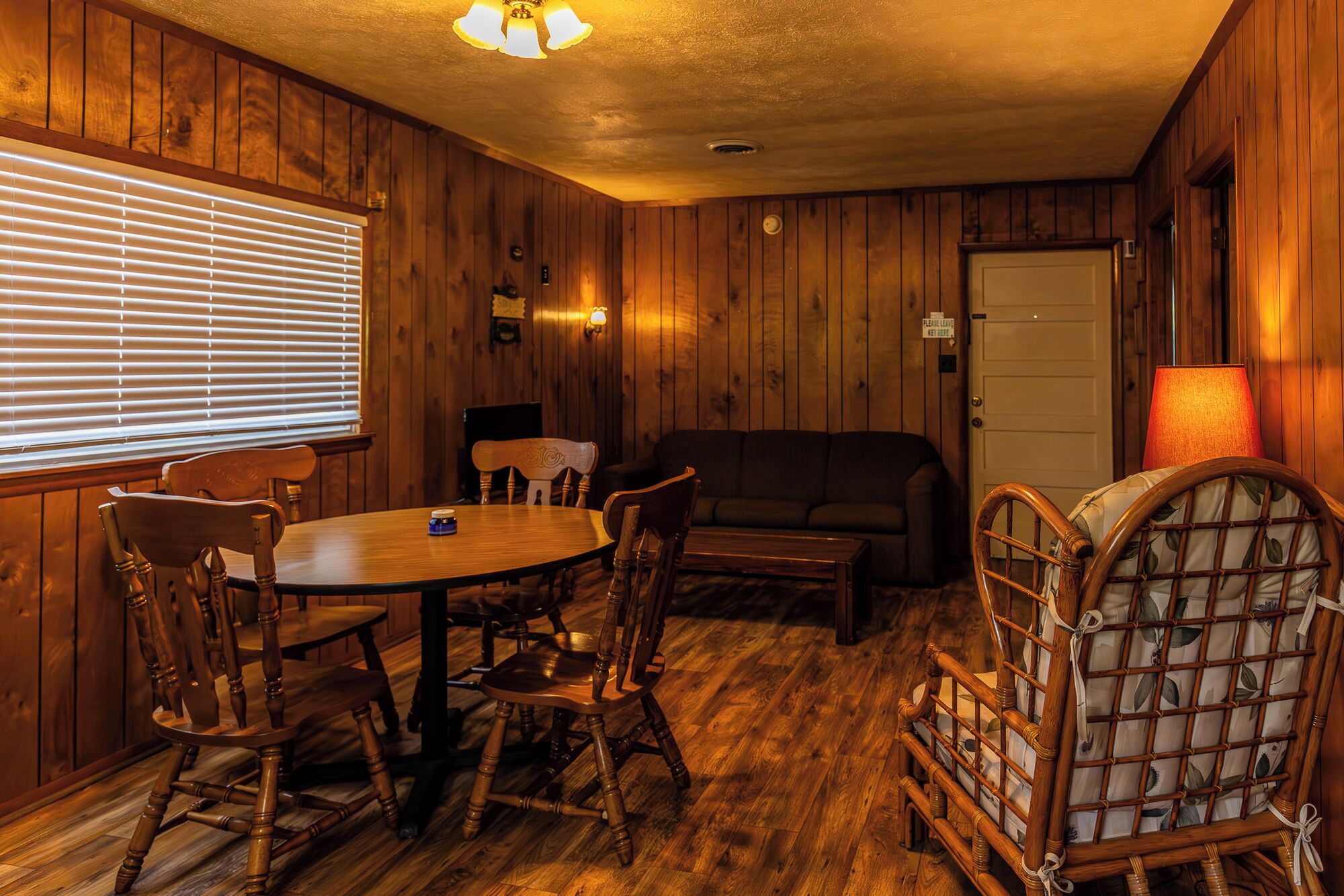 A room with wooden paneling, a round table with chairs, a sofa, and a lamp.