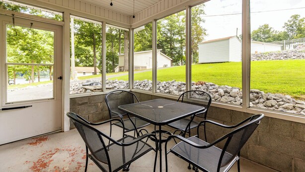 Een overdekt terras met een tafel en stoelen, grote ramen en uitzicht op een gazon en gebouwen.