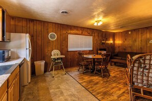 A compact kitchen with a refrigerator, microwave, and a small trash bin. There is a wooden dining table with chairs and a baby high chair. A window with blinds is visible, and a couch is in the background. - Peaceful Cabin on Kentucky Lake (Benton)