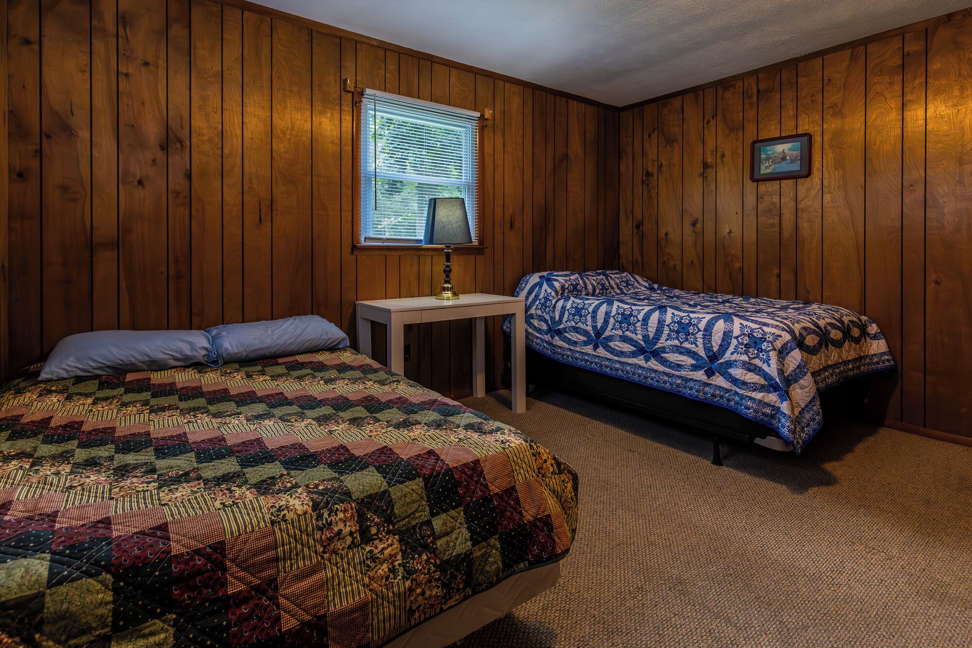 Une chambre avec des lambris en bois, un lit recouvert d’une courtepointe colorée, une table de chevet avec une lampe, et un lit superposé avec une courtepointe bleue et blanche.