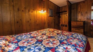 Una habitación con paredes de madera, un edredón acolchado, un aparador y una lámpara fijada a la pared.