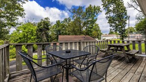 A wooden deck with metal chairs and tables, surrounded by a wooden railing, overlooking a grassy area with trees and houses in the background. - Spacious Cabin on Kentucky Lake (Benton)
