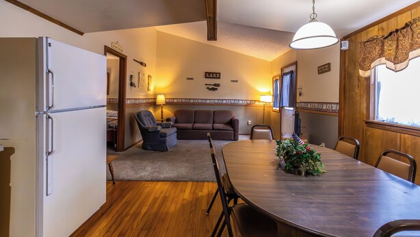 A compact kitchen with a refrigerator, dining table, and chairs. A sofa and armchair are in the adjacent living area. - Spacious Cabin on Kentucky Lake (Benton)