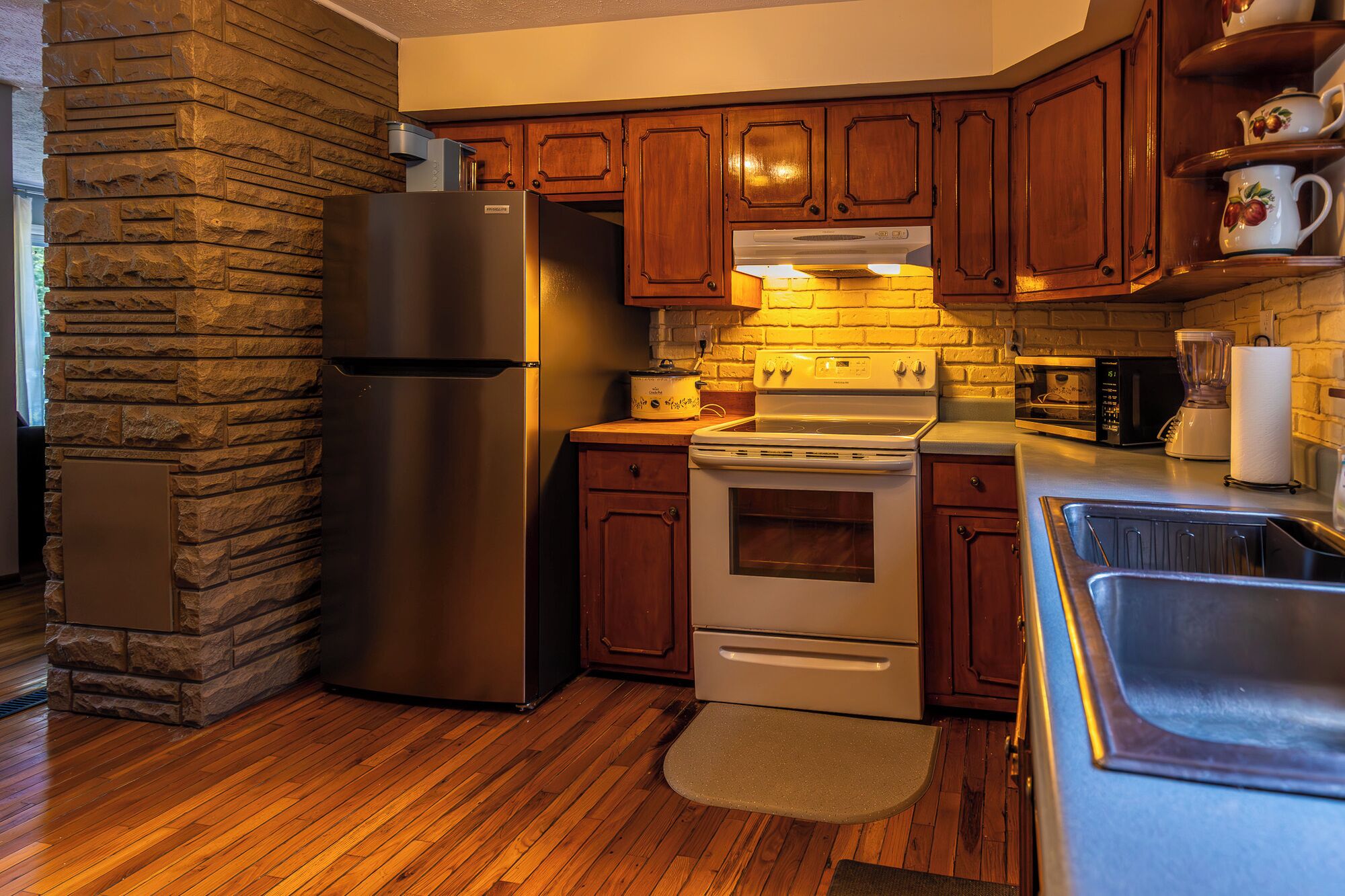 Una cocina con detalles en pared de piedra, armarios de madera, refrigerador de acero inoxidable, estufa y microondas.