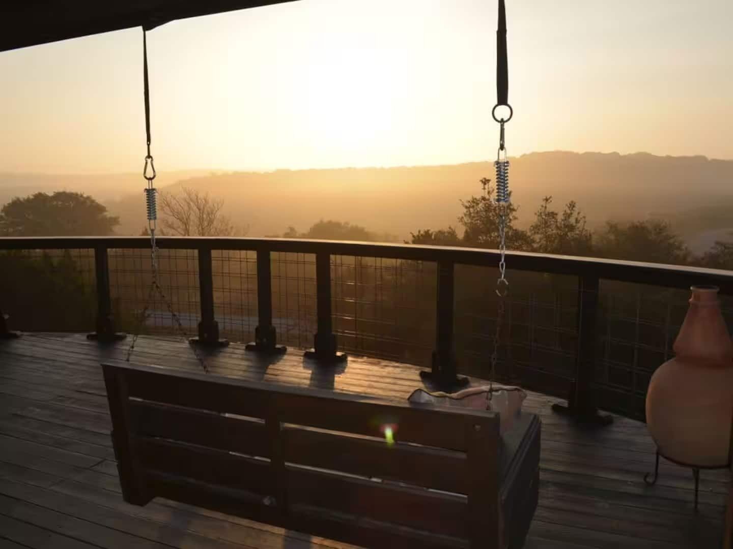 A wooden deck with a bench and a hanging chair, overlooking a landscape with trees and hills during sunset.