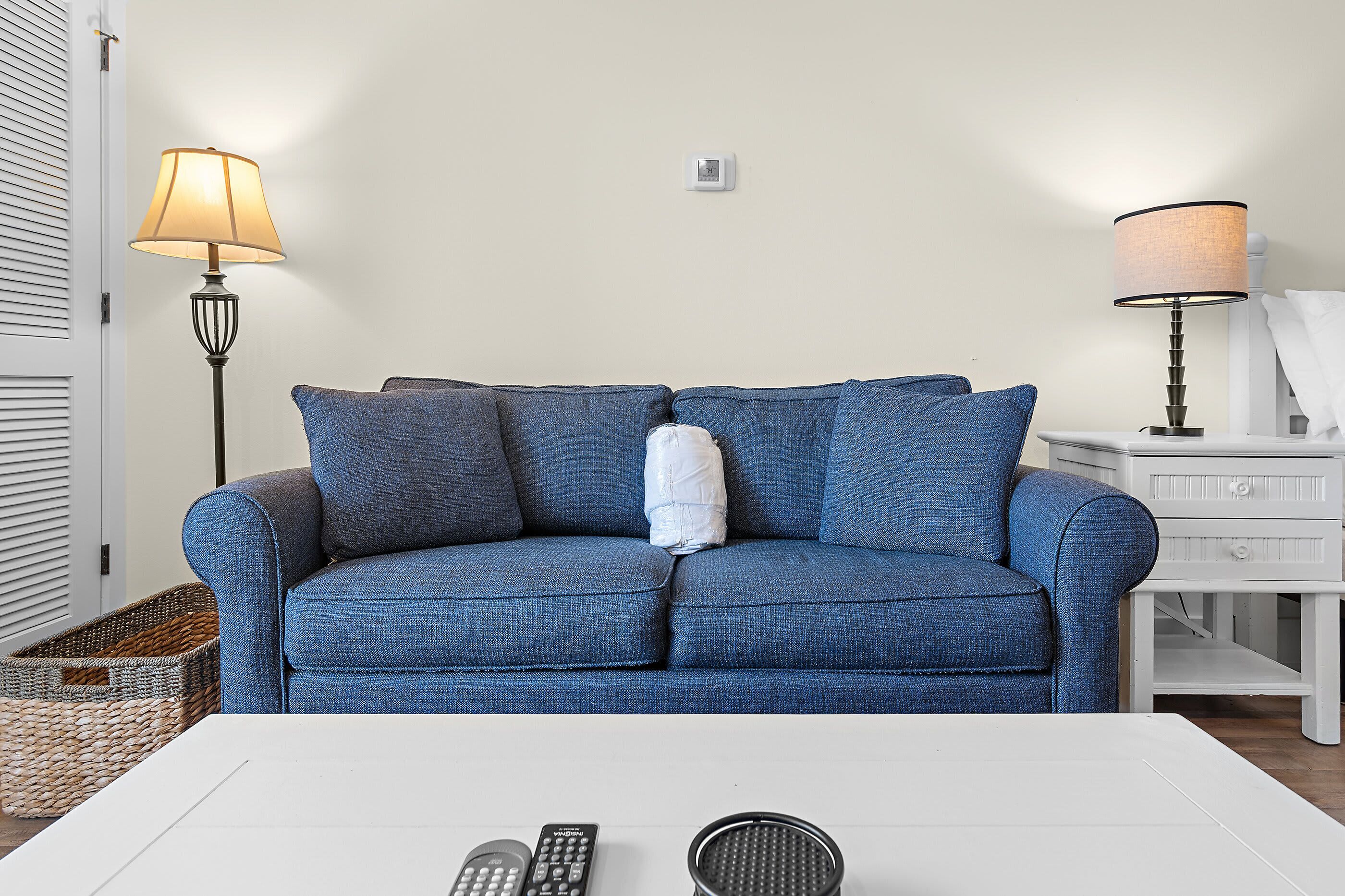 Un canapé bleu avec un coussin blanc, une lampe, une télécommande et un panier.