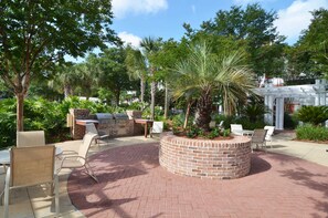 Un patio al aire libre con suelo de ladrillo, una jardinera circular de ladrillo y una zona de barbacoa rodeada de sillas y mesas blancas.