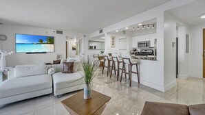 A modern living room with a white sofa, a coffee table, and a TV displaying a beach scene. A kitchen with white cabinets and a dining area with chairs are visible in the background. - Luxurious private unit with parking (Miami)