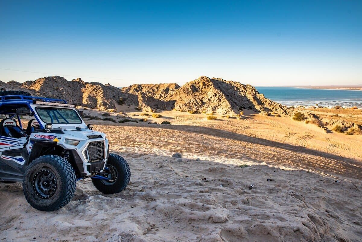 A white off-road vehicle parked on a sandy beach with rocky hills in the background.