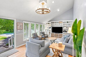 A modern living room with a television, seating, and a large window with blinds. - Elegant retreat, 10 min. from Newport; game room, fire pit, & huge deck! (Jamestown)
