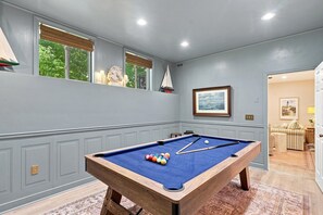 Mesa de bilhar em um quarto com vista para o verde através de janelas, um quadro emoldurado na parede e uma porta que dá para outro cômodo.
