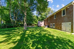 A residential property with a well-maintained lawn, a wooden house, and a large tree providing shade. - Elegant retreat, 10 min. from Newport; game room, fire pit, & huge deck! (Jamestown)