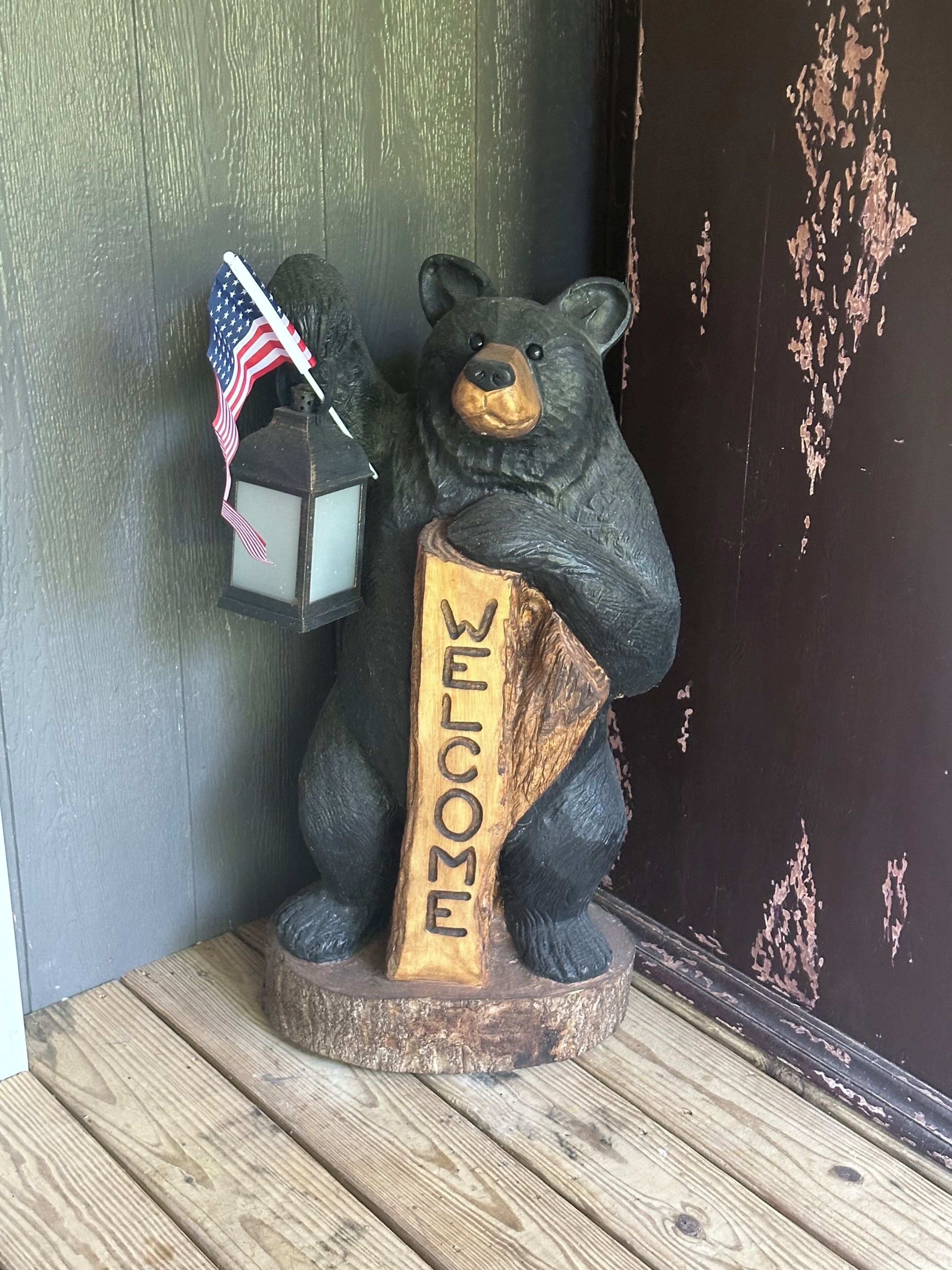 Een houten beerbeeld met 'WELCOME' in een boomstomp gekerfd, een lantaarn met een Amerikaanse vlag, en een donkere houten achtergrond.