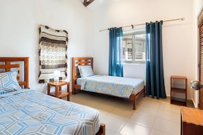 A room with two beds, a window with curtains, a wooden headboard, a nightstand, and a small shelf. - Hacienda Iguana 3BR Golf & Pool View Condo (Tola)