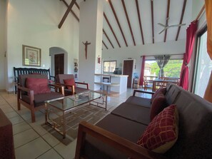 A spacious living area with a sofa, chairs, and a glass coffee table. A kitchen area is visible in the background. - Hacienda Iguana 3BR Golf & Pool View Condo (Tola)
