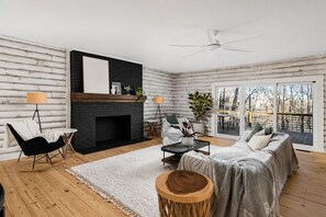 Ein Blockhaushaus-Interieur mit Kamin, Deckenventilator, einer Couch, einem Sessel und Blick ins Freie.