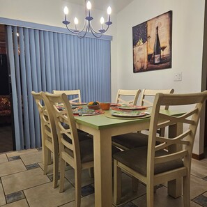 Un comedor con una mesa dispuesta para cuatro personas, rodeada de sillas, una araña y un cuadro de una botella de vino en la pared.