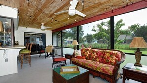 A spacious room with a wooden ceiling, a large window, a floral sofa, a wooden coffee table, and a dining area with chairs and a table. - Coastal Comfort Retreat - Pet-Friendly Paradise w/ Fenced Yard, Fire Pit & Lanai (Fort Pierce)