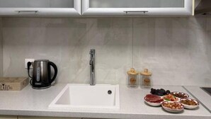 A kitchen with light-colored cabinets, a white sink, a kettle, and a variety of food items on the counter. - Le Grace Urban Retreat Near Near Citycenter Sea (Kalamaria)