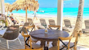 Um restaurante à beira-mar com cadeiras de vime e uma mesa redonda de madeira, com vista para uma praia arenosa e águas azul-turquesa.