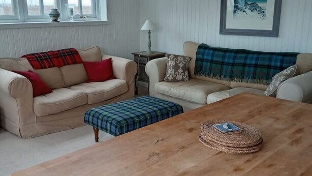 A living room with two beige sofas, one with a plaid blanket and red pillows, and the other with a blue blanket and patterned pillows. A wooden coffee table with a woven basket and a book is in the center.