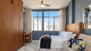 Quarto de hotel com cama, escrivaninha, cadeira, ventoinha no teto e janela com vista para a cidade.