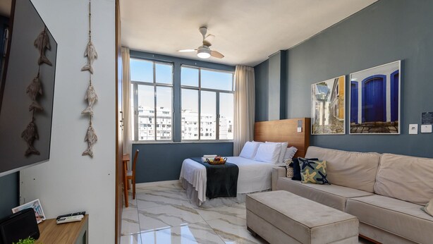 Quarto de hotel com cama, sofá, escrivaninha com computador, ventilador de teto e janelas amplas.