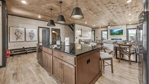 A modern kitchen with wooden cabinets, a central island, pendant lighting, and a dining area with wooden chairs.