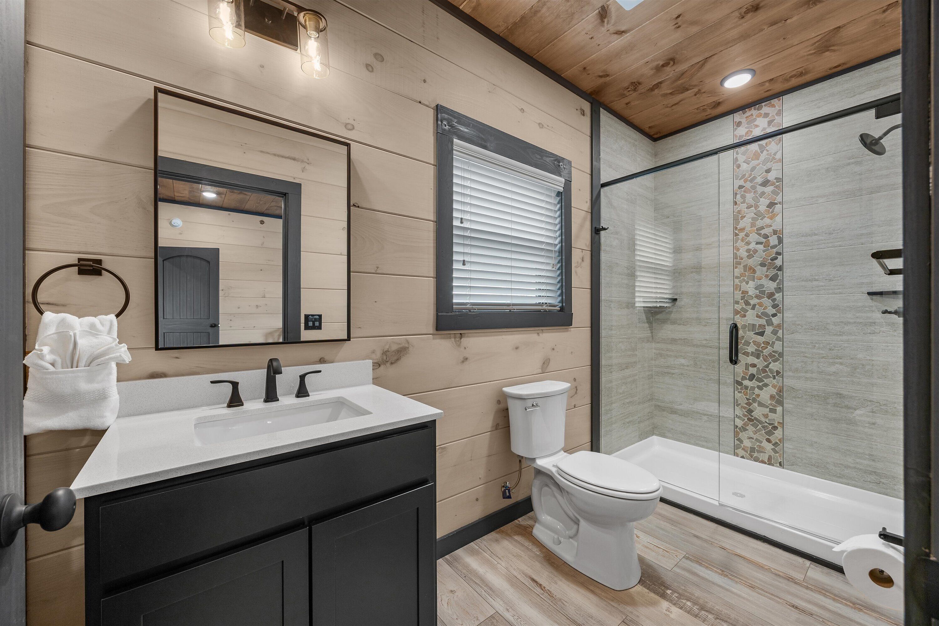 Een moderne badkamer met een houten wand, een grote spiegel, een wit wastafelmeubel, toilet en een douchecabine met glazen wanden.