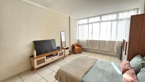 Un dormitorio con una cama, un televisor sobre un soporte de madera, un taburete pequeño, un espejo y una ventana con cortinas.