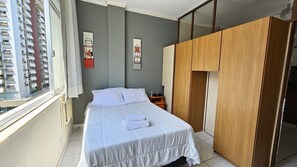 A bedroom with a bed, a wooden wardrobe, and a window with a view of a building. - Studio Zen Copacabana w/ AC 5 min to Beach (Rio de Janeiro)