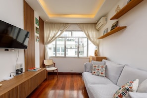 Uma sala de estar moderna com uma televisão de tela plana, painéis de madeira, um sofá com almofadas estampadas, uma poltrona e uma janela com cortinas.
