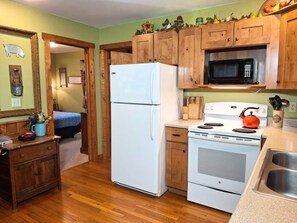 A compact kitchen with wooden cabinets, a white refrigerator, a microwave, and a stove. There is a small sink and a countertop with a kettle. - Old Man's Haven - Cabin 1 (Logan)