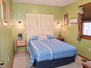 Un dormitorio con cama, dos mesitas de noche, una mesita auxiliar, una ventana con cortinas y una lámpara de pared.
