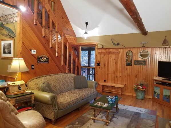 Interior acogedor de una cabaña con escalera de madera, un sofá, una mesa de centro y un televisor.