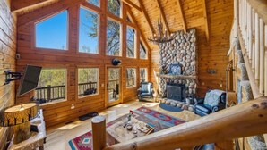 Interno di una baita di legno con camino in pietra, ampie finestre e una scala in legno.