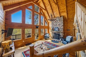 A log cabin interior with a stone fireplace, large windows, and a wooden staircase. - Mountain View Log Cabin w Hot Tub & Foosball (Bryson City)