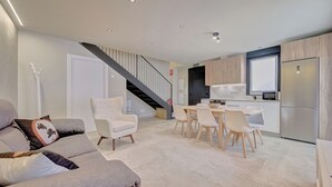 A modern living room with a grey sofa, white armchairs, and a dining area with a table and chairs. A kitchen with a fridge, stove, and cabinets is visible in the background. - LA FRAGUA ANCÍN APARTMENT (Ancín)