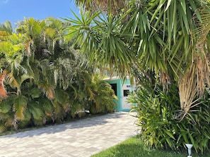 Un chemin pavé mène à un bâtiment doté d’une porte bleue, qui est entouré d’une végétation tropicale luxuriante.