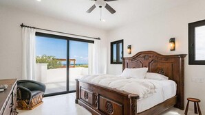 Een slaapkamer met een groot bed, een plafondventilator, een bureau met stoel en uitzicht op de omgeving door een schuifdeur.