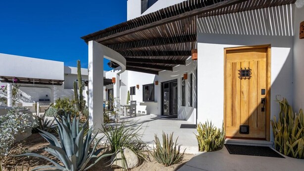 Een modern gebouw met een houten deur, witte muren en een overdekt terras met cactussen en planten.