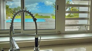 A kitchen sink with a view of a swimming pool and a cloudy sky through a window.
