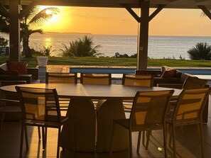 Un área de comedor al aire libre con vista al océano al atardecer, que incluye una mesa redonda con sillas, una piscina y un espacio de estar cubierto con cojines.