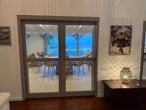 Una habitación con puerta corrediza de vidrio que da a un patio con vista al océano. Hay un comedor con sillas y una mesa, y una lámpara decorativa sobre una mesita auxiliar.