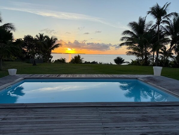 Una piscina rectangular con cubierta de madera, rodeada de palmeras y vistas a la puesta de sol sobre el océano.