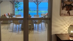 A room with a view of the ocean through a glass door, a dining area with chairs and a table, and a decorative vase on a side table.