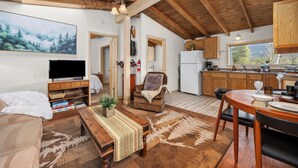 A cozy living room with a TV, sofa, and a fireplace. There is a dining area with a table and chairs. The kitchen has wooden cabinets and a window.