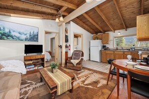 A cozy living room with a TV, sofa, and a fireplace. There is a dining area with a table and chairs. The kitchen has wooden cabinets and a window.