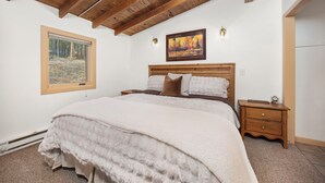 A bedroom with a wooden ceiling, a large bed with a fluffy white blanket, a nightstand with a clock, and a painting on the wall.